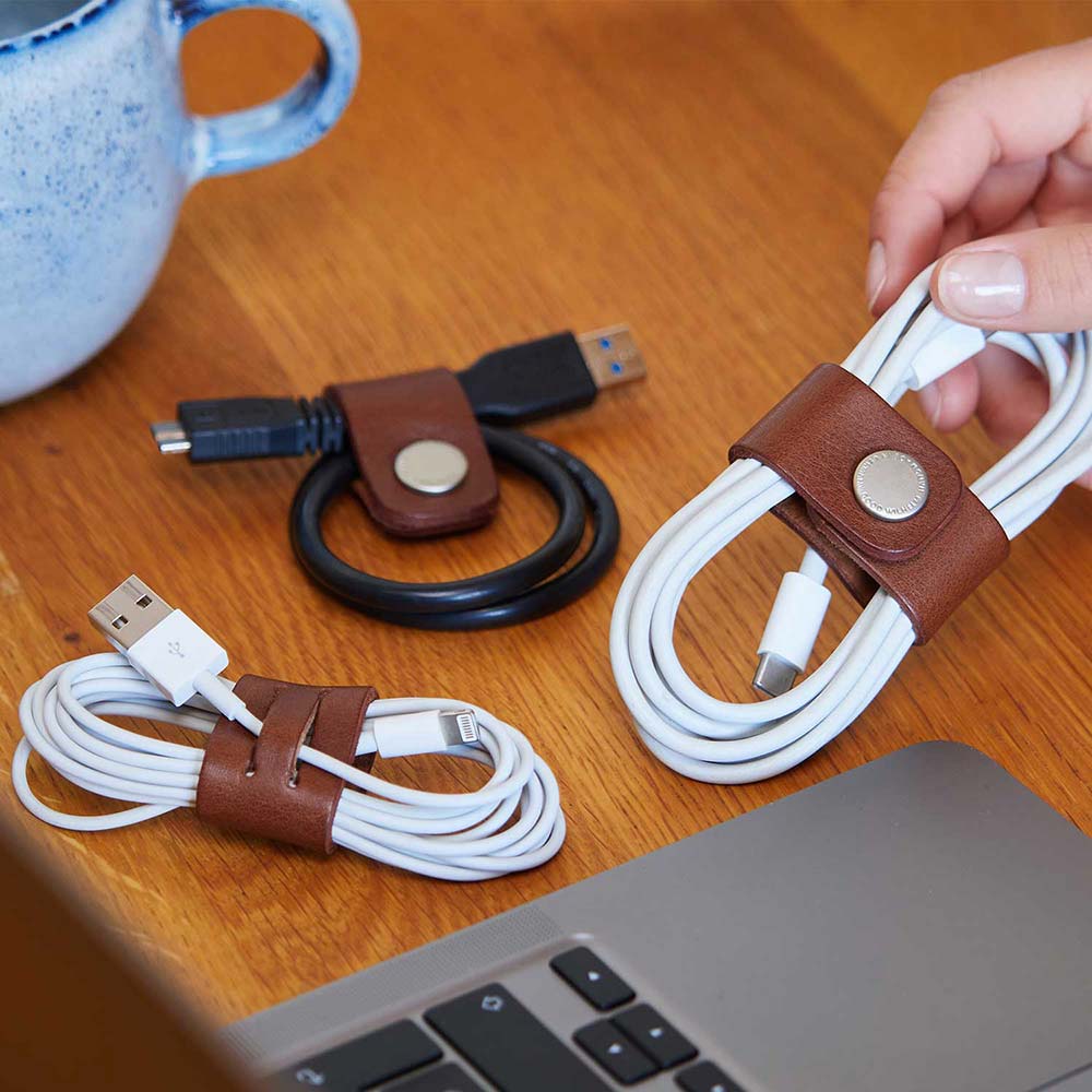 Three computer cables with leather cable organizers in the color Cognac from Good Wilhelm lie on a wooden table
