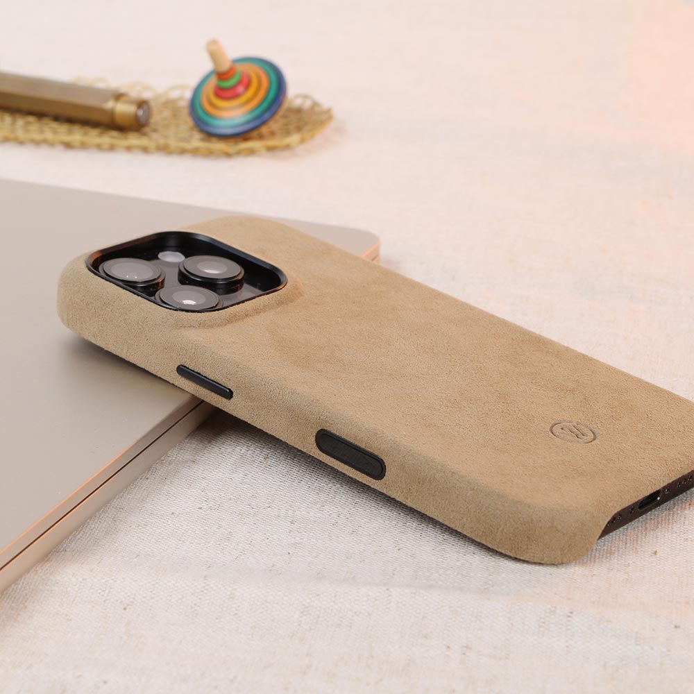 Beigefarbene Alcantara-Hülle für iPhone 16 Pro von Good Wilhelm, handgefertigt mit integriertem Kamera-Kontrollknopf, stilvoll dargestellt auf einem Tisch mit Accessoires.