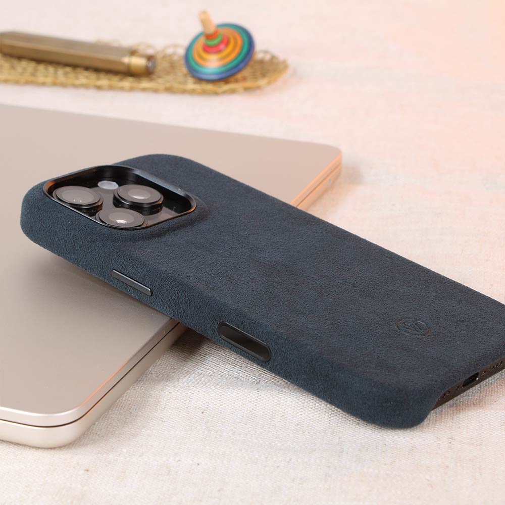 Blaue Alcantara-Hülle für iPhone 16 Pro von Good Wilhelm, handgefertigt mit integriertem Kamera-Kontrollknopf, geschmackvoll platziert auf einem Tisch mit Accessoires.