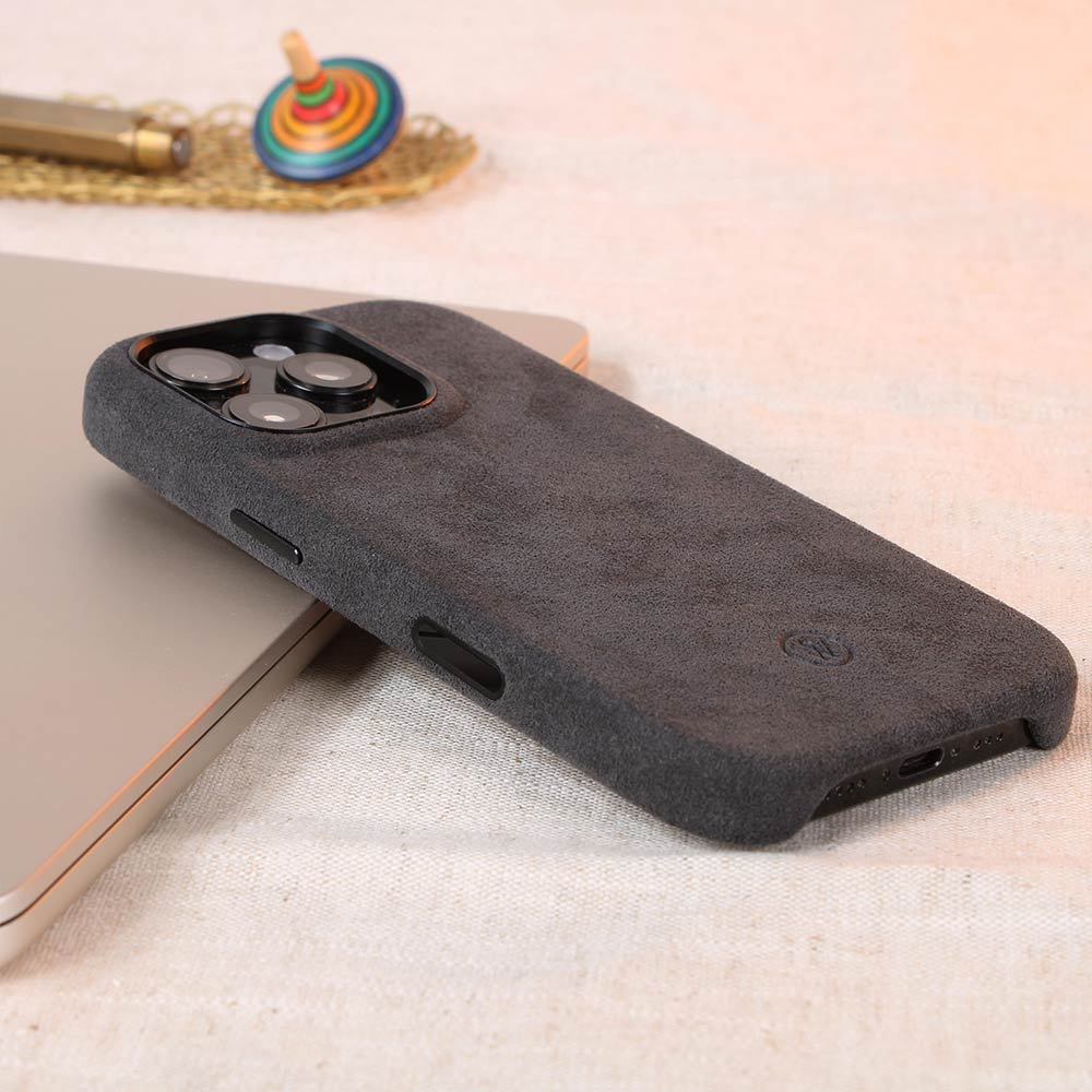Dunkelgraue Alcantara-Hülle für iPhone 16 Pro von Good Wilhelm, handgefertigt mit integriertem Kamera-Kontrollknopf, elegant präsentiert auf einem Tisch mit Accessoires.