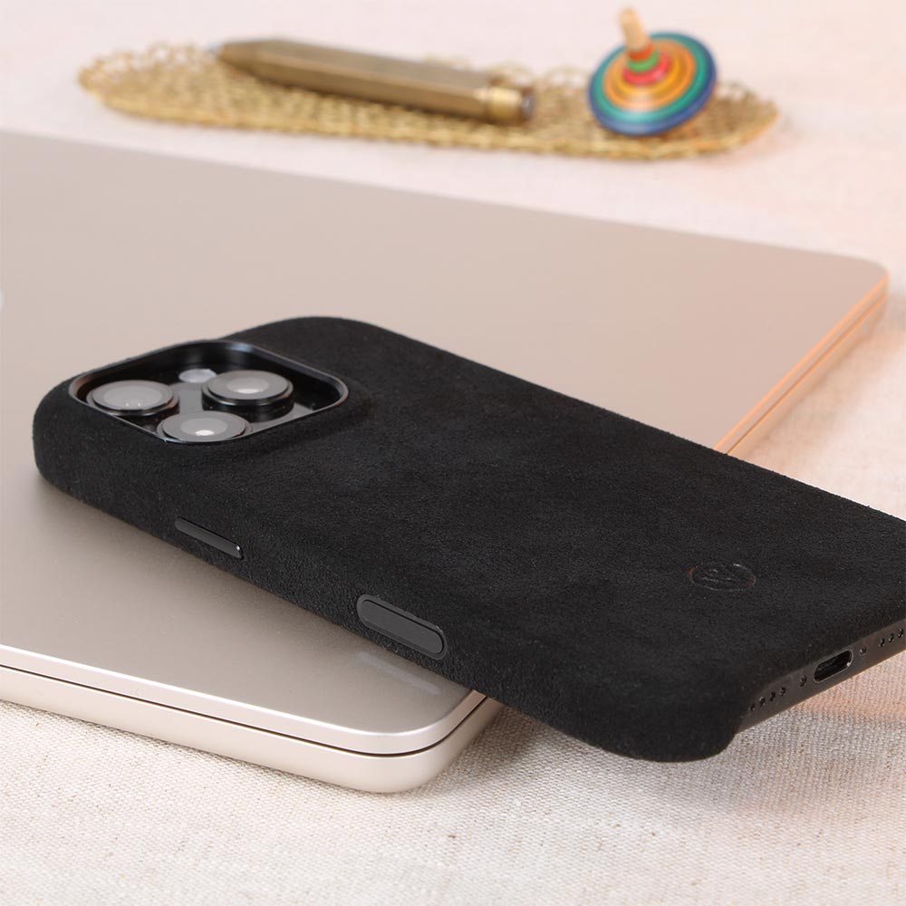 Handgefertigte schwarze Alcantara-Hülle für iPhone 16 Pro von Good Wilhelm mit integriertem Kamera-Kontrollknopf, stilvoll präsentiert auf einem Tisch mit Accessoires.