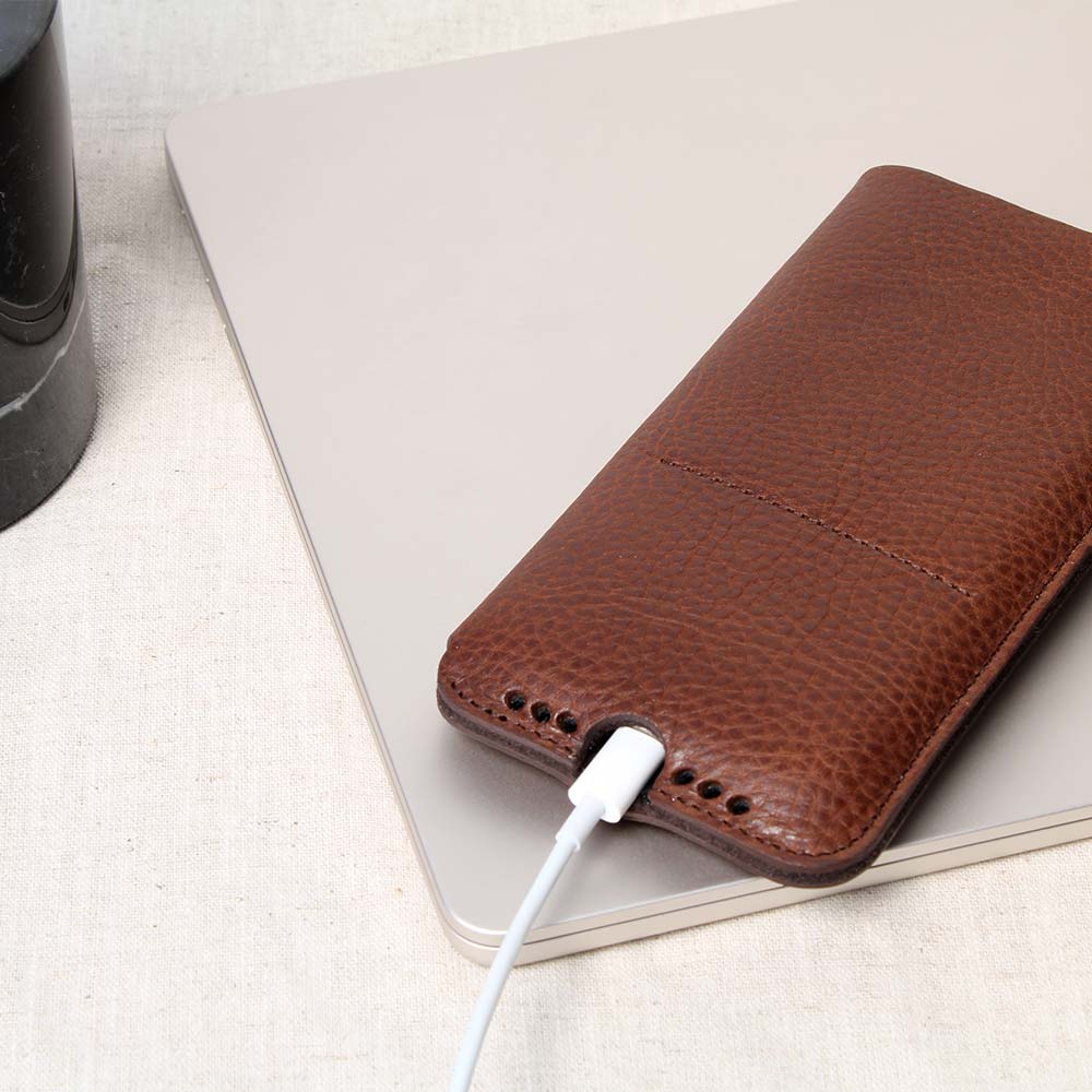 Brown iPhone sleeve from Good Wilhelm, made of high-quality leather with charging recess and soundholes, based on a MacBook on a table - elegant protective case made of fine leather for optimum protection and functionality