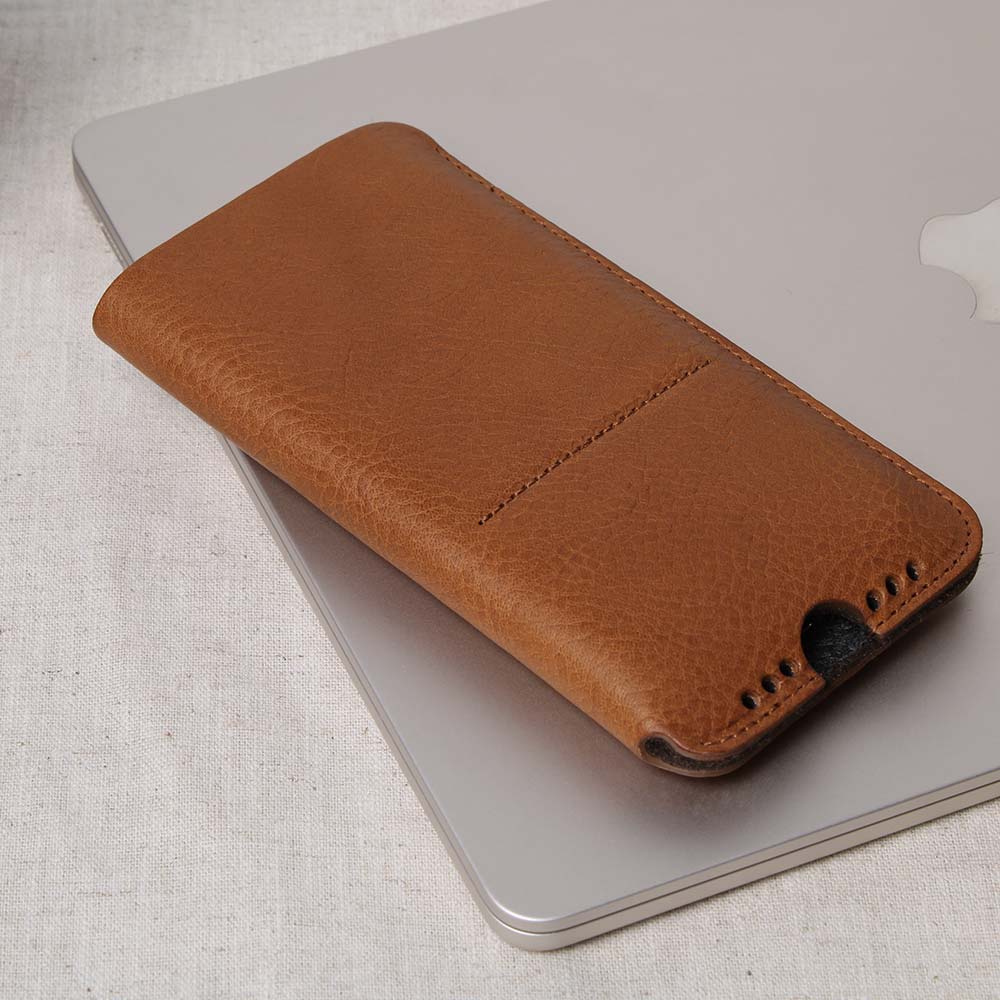 Cognac-brown iPhone sleeve made of high-quality leather with charging cut-out and soundholes, based on a MacBook on a table - stylish protective case made of fine leather for optimum protection and functionality.