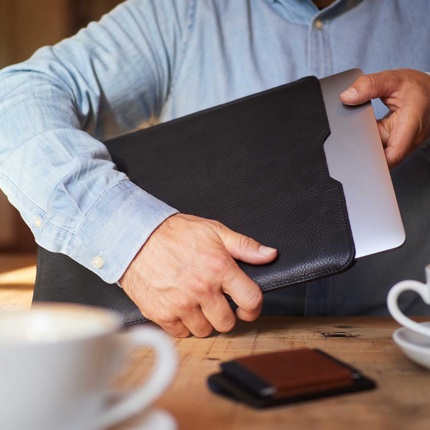 Ein Mann an einem Holztisch zieht ein Macbook aus einer schwarzen Lederhülle von GoodWilhelm